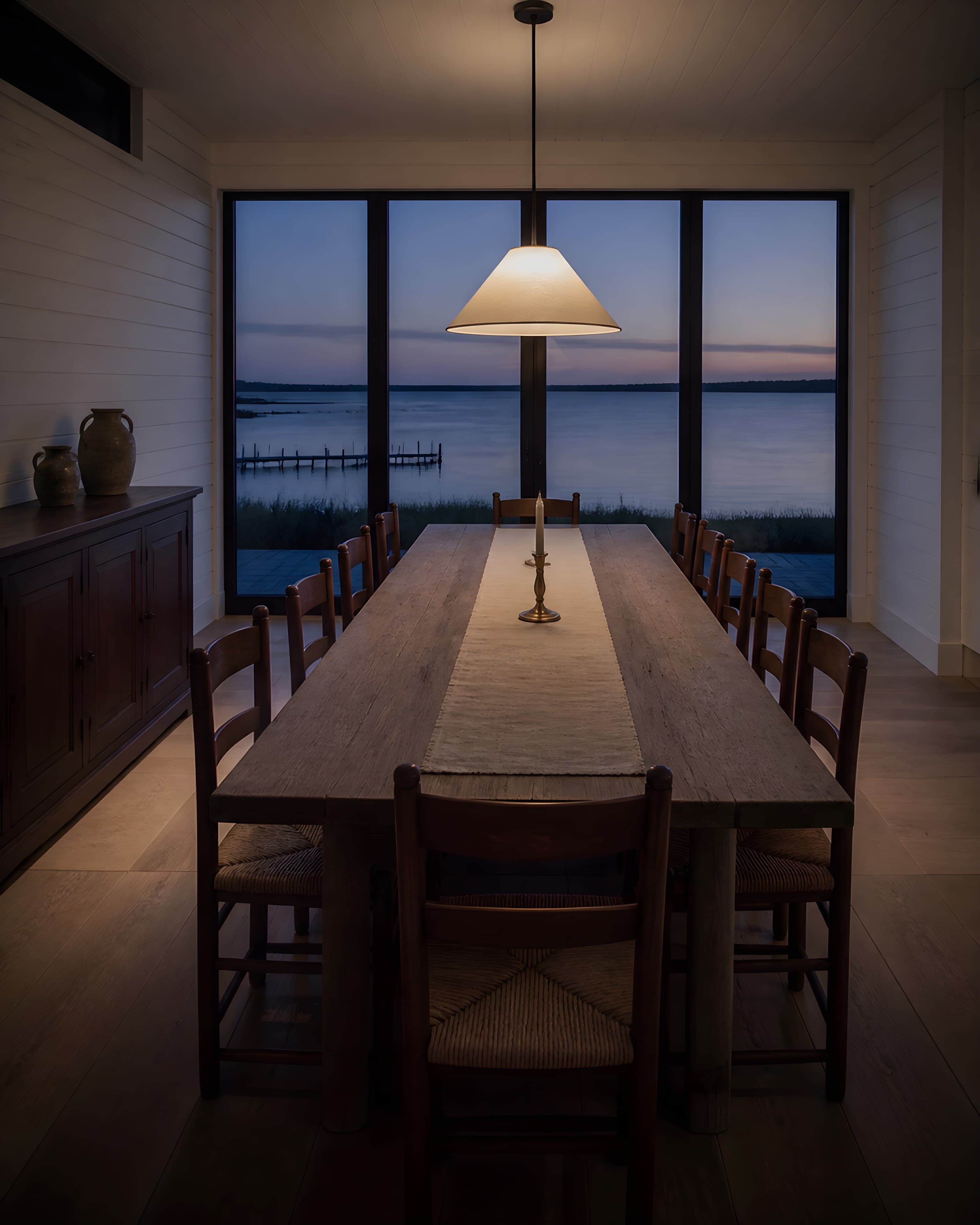 The dining room at dusk, a long oak table under a linen pendant with the last light on the bay through tall casements